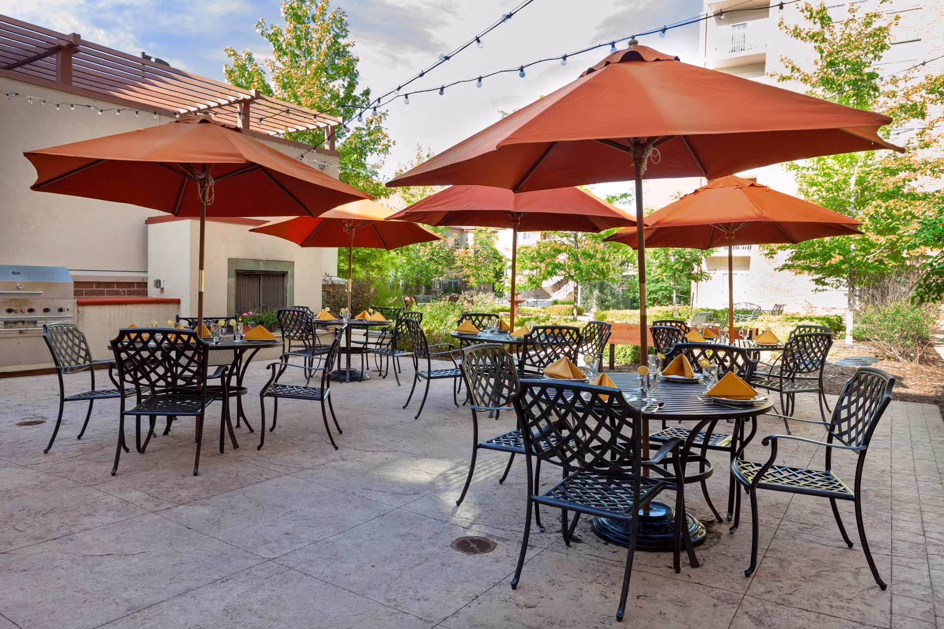 Outdoor patio area with several black metal tables and chairs, each table shaded by large orange umbrellas. Tables are set with yellow napkins and glassware. There is a built-in grill and a fireplace on the left side, surrounded by greenery and trees under a partly cloudy sky.