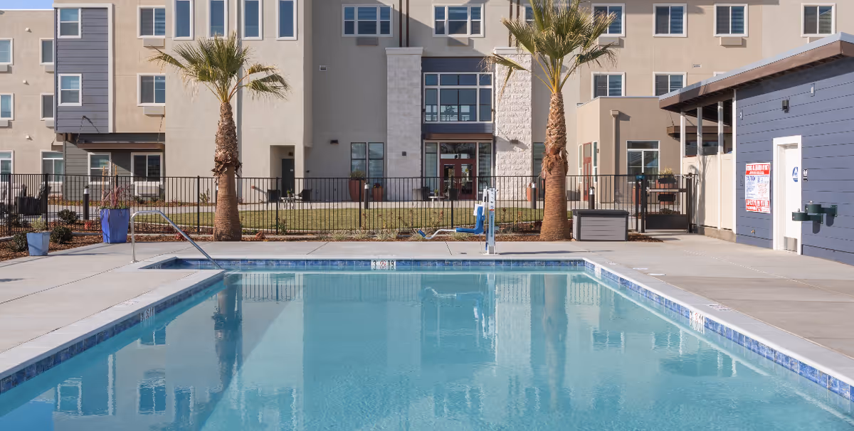 Outdoor swimming pool with clear blue water in front of a multi-story residential building. Two palm trees are planted near the pool area, which is surrounded by a concrete deck and a black metal fence. There is a pool lift for accessibility and a blue building with a water fountain and safety signs on the right side.
