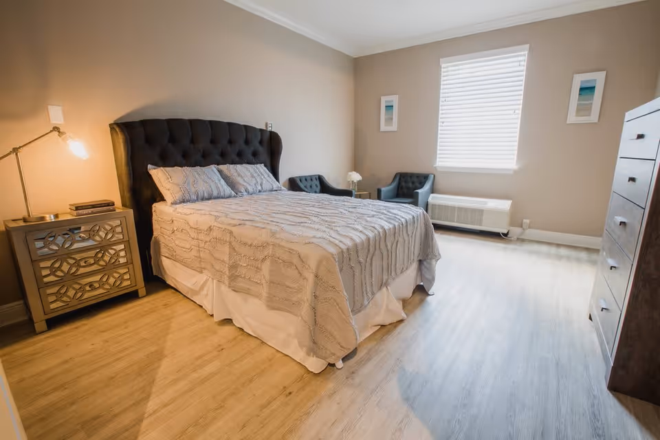 Well-lit bedroom with a tufted headboard bed, nightstand with lamp, two armchairs by a window, and a dresser.