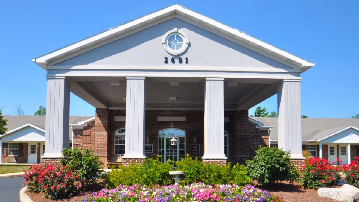 Front exterior view of a senior living facility building with a large covered entrance supported by four white columns. The building has a brick facade with white trim and a circular window above the entrance displaying the number 2601. There are well-maintained flower beds with colorful flowers and green shrubs in front of the building under a clear blue sky.