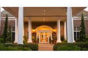 Entrance of a building with large white columns, glass double doors, and warm lighting inside. The building is surrounded by neatly trimmed bushes and small trees.