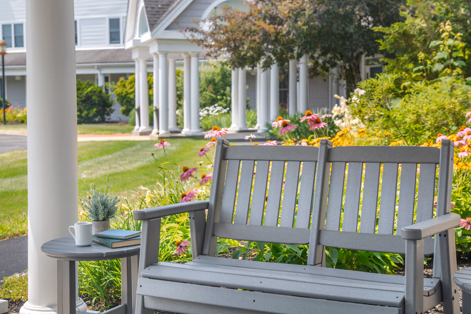 A gray outdoor bench and side table with a white mug, two books, and a small potted plant on it, set in a garden area with colorful flowers and greenery. In the background, there is a building with white columns and a porch.
