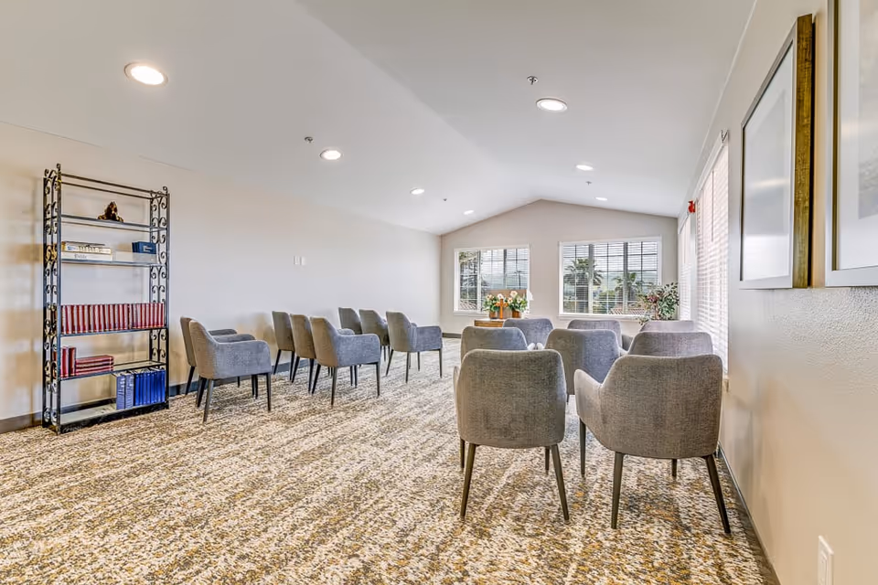 A bright, spacious room with beige walls and patterned carpet featuring two rows of gray upholstered chairs facing a window with blinds. A black metal bookshelf with books and decorative items is positioned against the left wall. The room is well-lit with recessed ceiling lights and has framed artwork on the right wall.