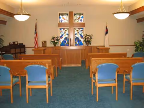 Wood pews face a cross-shaped stained glass window behind a pulpit in a chapel.