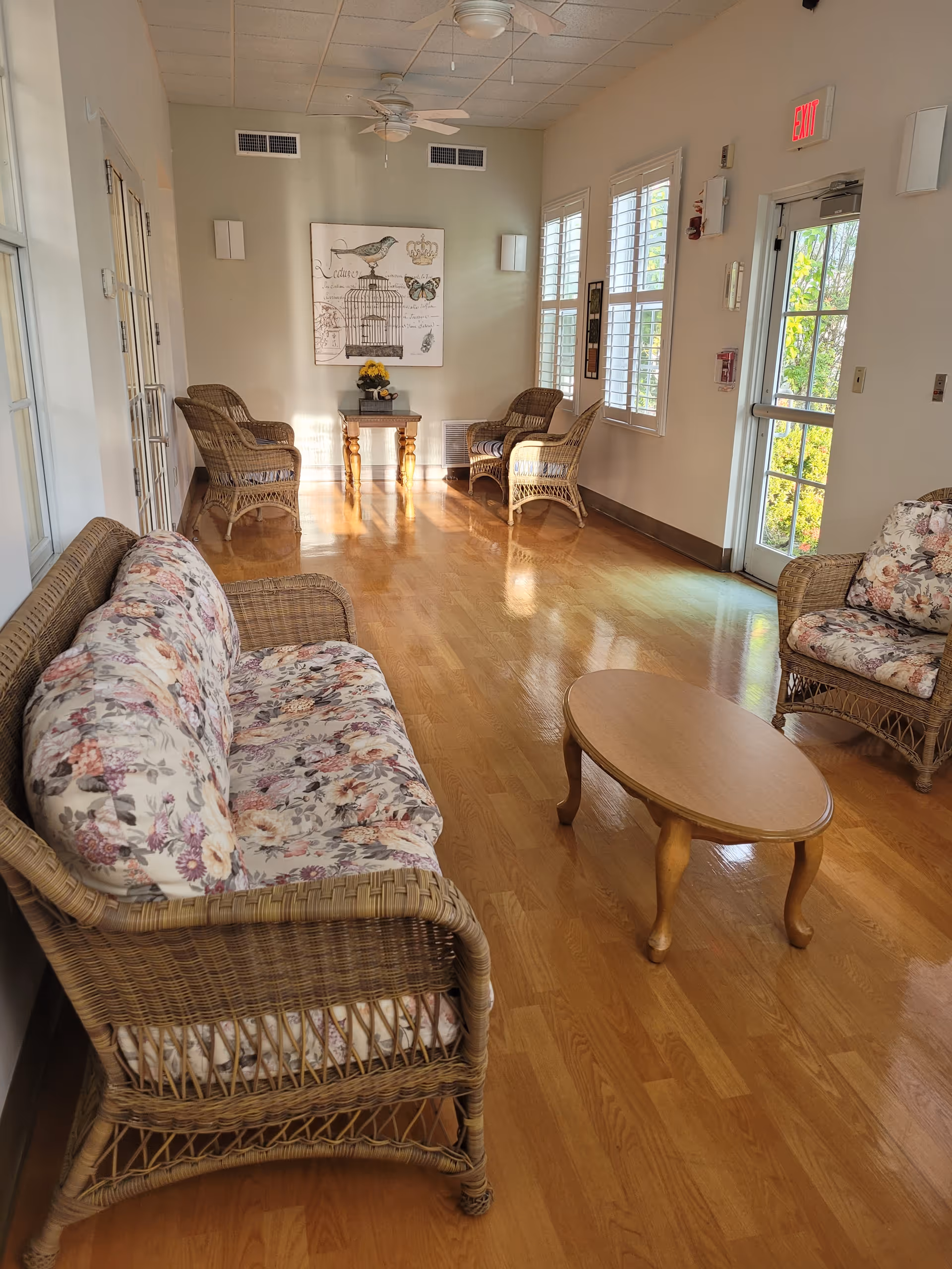A bright, cozy sitting area with wicker furniture including a floral cushioned sofa, two armchairs, and a wooden coffee table on a polished wooden floor. The room has large windows with white shutters, a ceiling fan, and a wall decoration featuring a bird and a birdcage. An exit door with glass panels is visible on the right side.