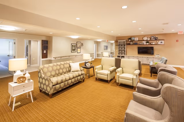 A spacious and well-lit living room area in a senior living facility featuring a patterned sofa, several armchairs arranged in a semi-circle, side tables with lamps, and a wall-mounted television with shelves and decorative items above a long cabinet.