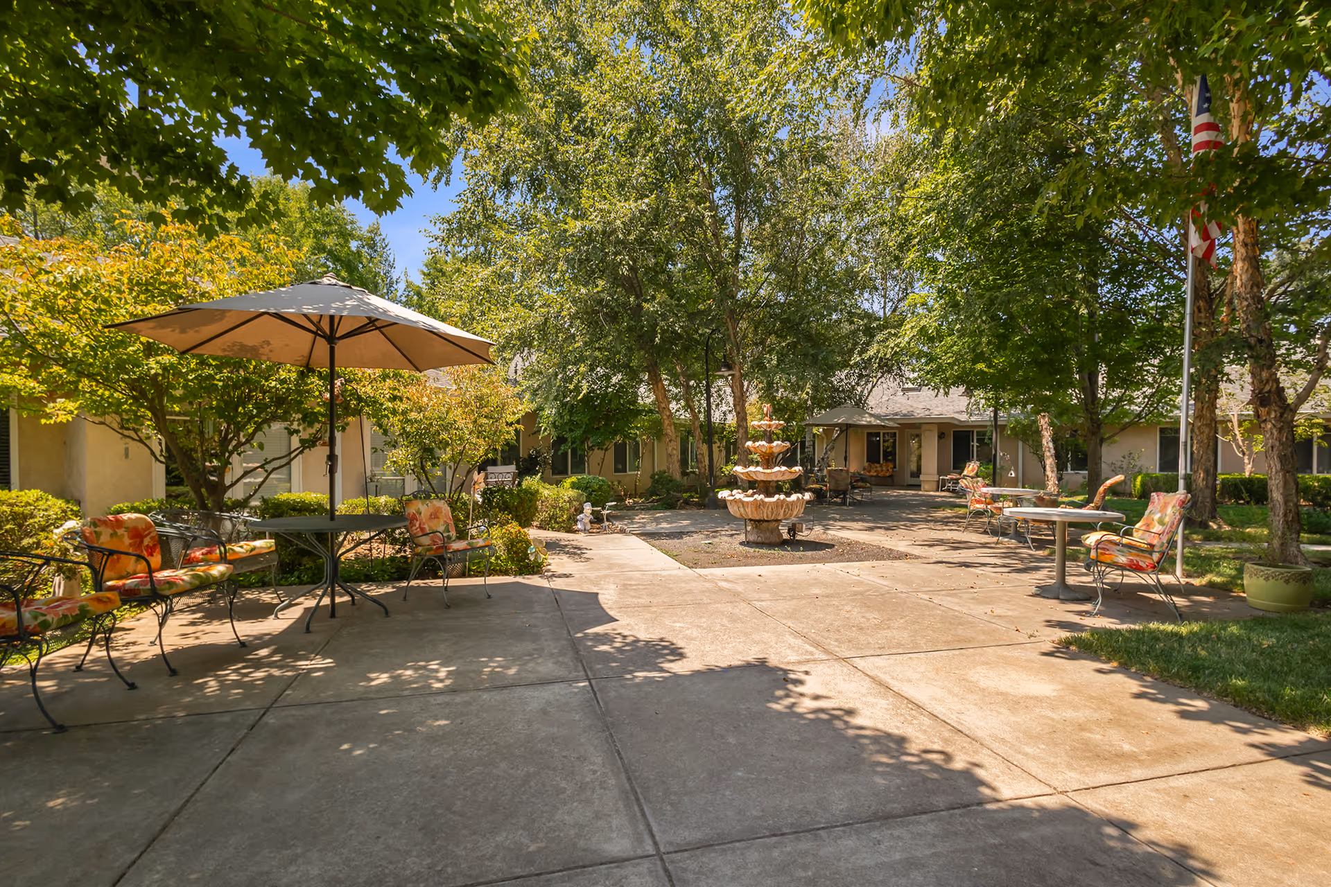 Outdoor courtyard area with concrete pathways, several tables with umbrellas and cushioned chairs, surrounded by trees and shrubs. A multi-tiered fountain is centered in the courtyard, and a building with a covered entrance is visible in the background under a clear blue sky.