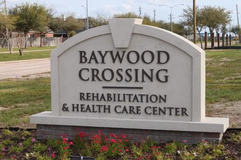 A large stone sign outdoors with the text 'Baywood Crossing Rehabilitation & Health Care Center' surrounded by grass, flowers, and trees in the background.