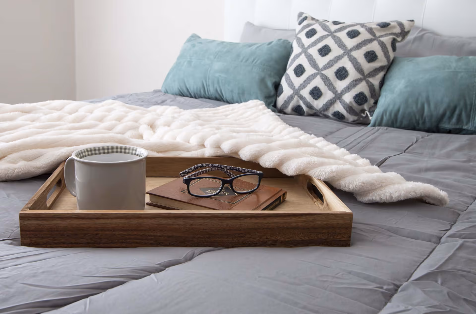 A cozy bed with a gray comforter, a white textured blanket, and decorative pillows in teal and gray patterns. On the bed is a wooden tray holding a white mug, a pair of eyeglasses, and two closed books.