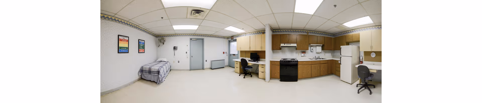 A spacious room in Hampton House featuring a single bed with a plaid blanket on the left side, a desk with a chair and computer in the middle, and a kitchen area on the right with wooden cabinets, a stove, a refrigerator, and a second desk with a chair. The room has white walls with a patterned border near the ceiling, a tiled ceiling with fluorescent lights, and a door at the back center.