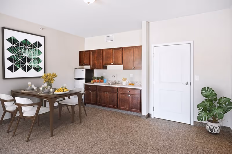 A bright and clean dining area with a wooden table set for four, featuring white cushioned chairs, a vase with yellow flowers, and a bowl of lemons. Behind the dining table is a kitchenette with dark wooden cabinets, a small refrigerator, a sink, and countertop with decorative items. A large framed geometric artwork with green tones hangs on the wall above the dining table. A white door and a potted green plant are visible on the right side of the room.