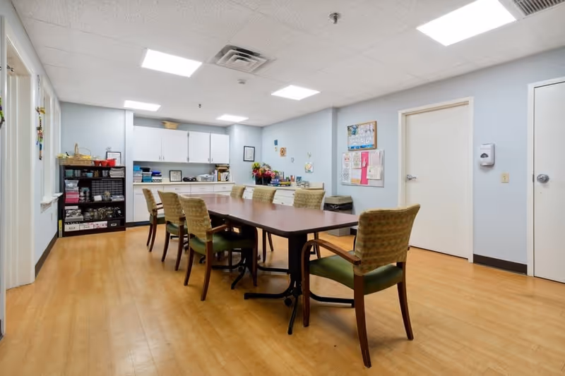 A well-lit room with a long rectangular table surrounded by eight cushioned chairs. The room has light blue walls, a wooden floor, and a white ceiling with recessed lighting. On one side, there are white cabinets and shelves with various items, including books and baskets. A bulletin board with papers and decorations is mounted on the wall. Two closed white doors are visible on the right side of the room.