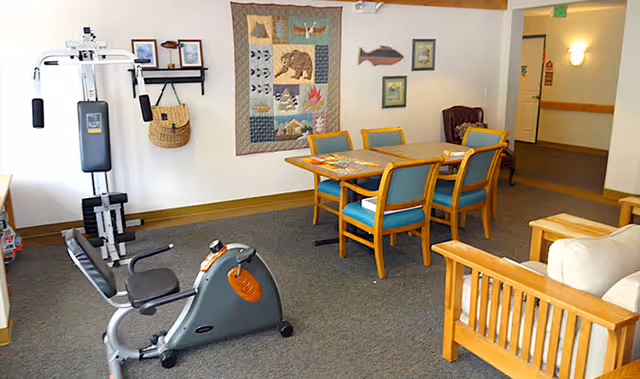A room in Mesa Vista Assisted Living featuring exercise equipment including a recumbent bike and a multi-function weight machine, a wooden table with four chairs, a wooden armchair with cushions, and wall decorations including a quilt with animal designs and framed pictures.