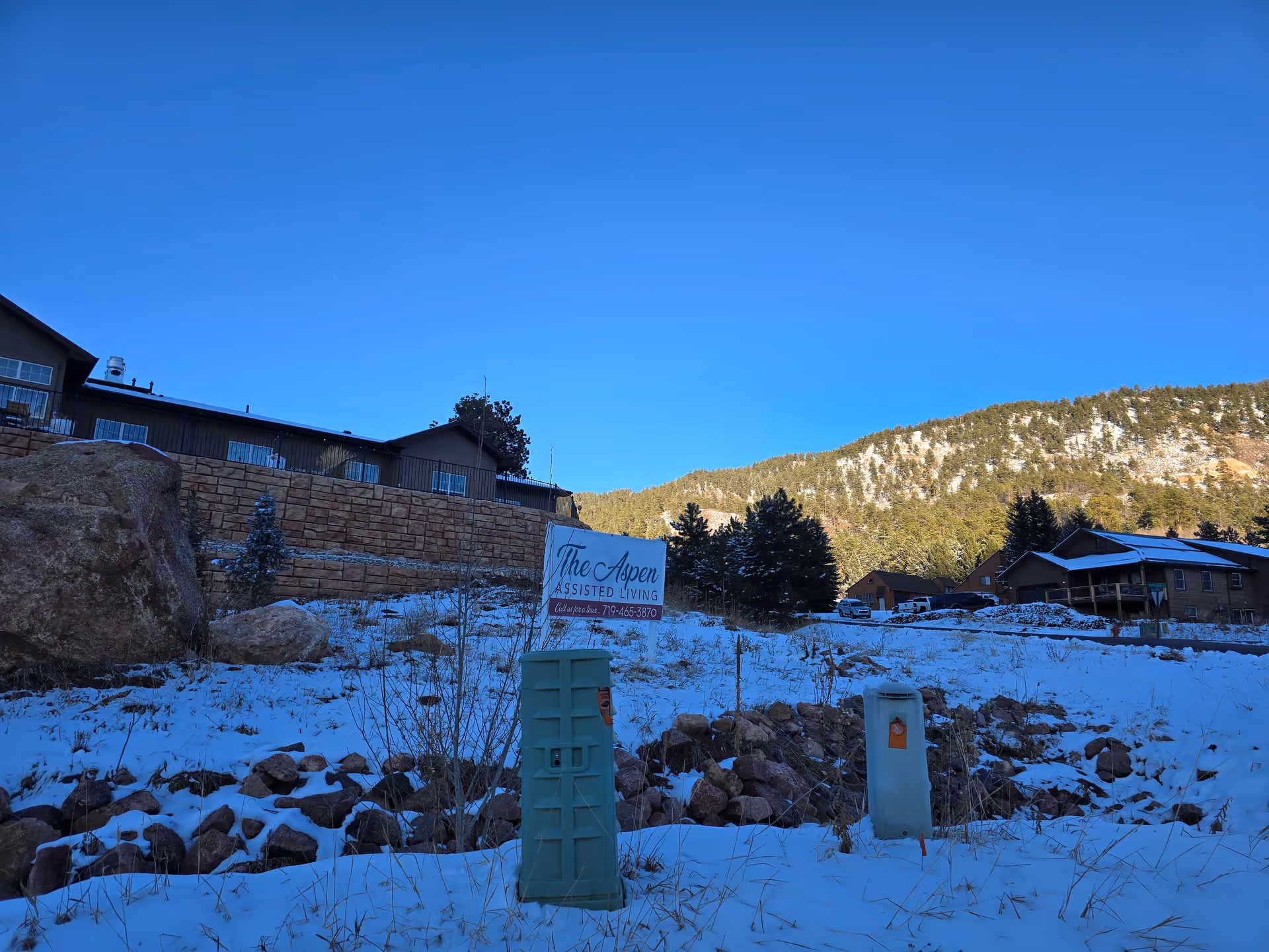 Snow-covered hillside with a sign that reads 'The Aspen Assisted Living' and the facility buildings against a forested mountain backdrop.
