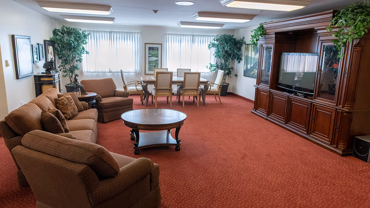 A spacious living room with brown upholstered sofas and armchairs arranged around a round wooden coffee table. In the background, there is a dining table with six chairs near large windows covered with sheer white curtains. The room features a large wooden entertainment center with a flat-screen TV and decorative plants on top. The floor is covered with a red carpet with a subtle pattern, and the walls are adorned with framed artwork and additional potted plants.