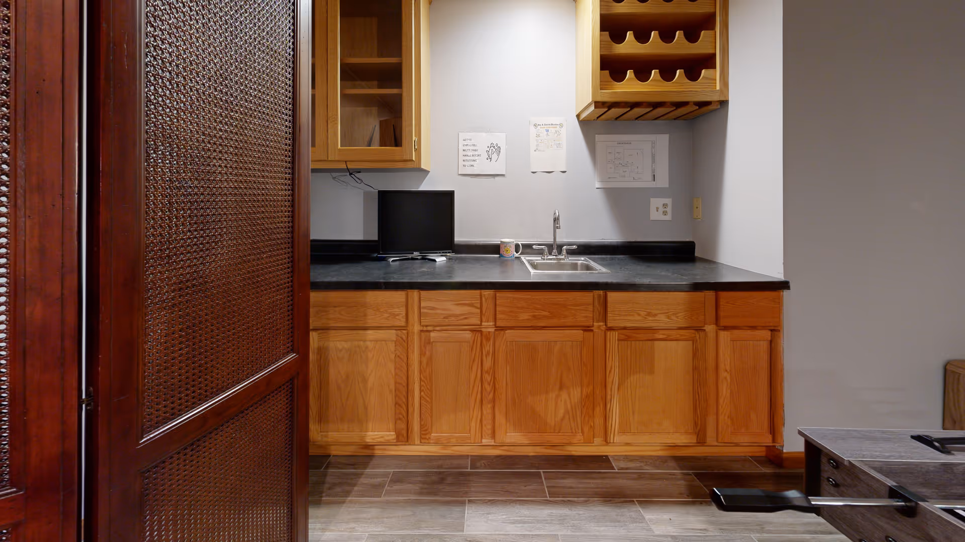 A small kitchen area with wooden cabinets, a black countertop, a stainless steel sink, and a mounted wooden wine rack. There is a small monitor and a coffee mug on the counter. The floor is tiled with large rectangular tiles, and a decorative wooden screen is partially visible on the left side.