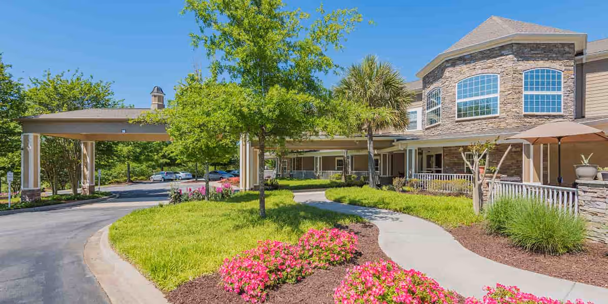 Exterior view of The Village at Southlake senior living facility showing a curved driveway, landscaped garden with pink flowers, green trees, and a two-story building with large windows and stone accents under a clear blue sky.