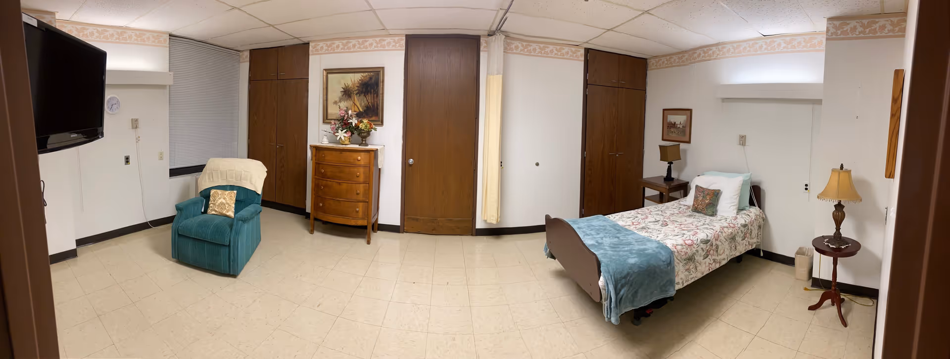 A senior living facility bedroom with a single bed covered in floral bedding and a blue blanket. There is a wooden nightstand with a lamp next to the bed, a small round table with another lamp, a wooden dresser with a floral arrangement on top, a teal armchair with a pillow and a blanket draped over it, a wall-mounted TV, and two wooden closets. The room has tiled flooring and white walls with a decorative border near the ceiling.