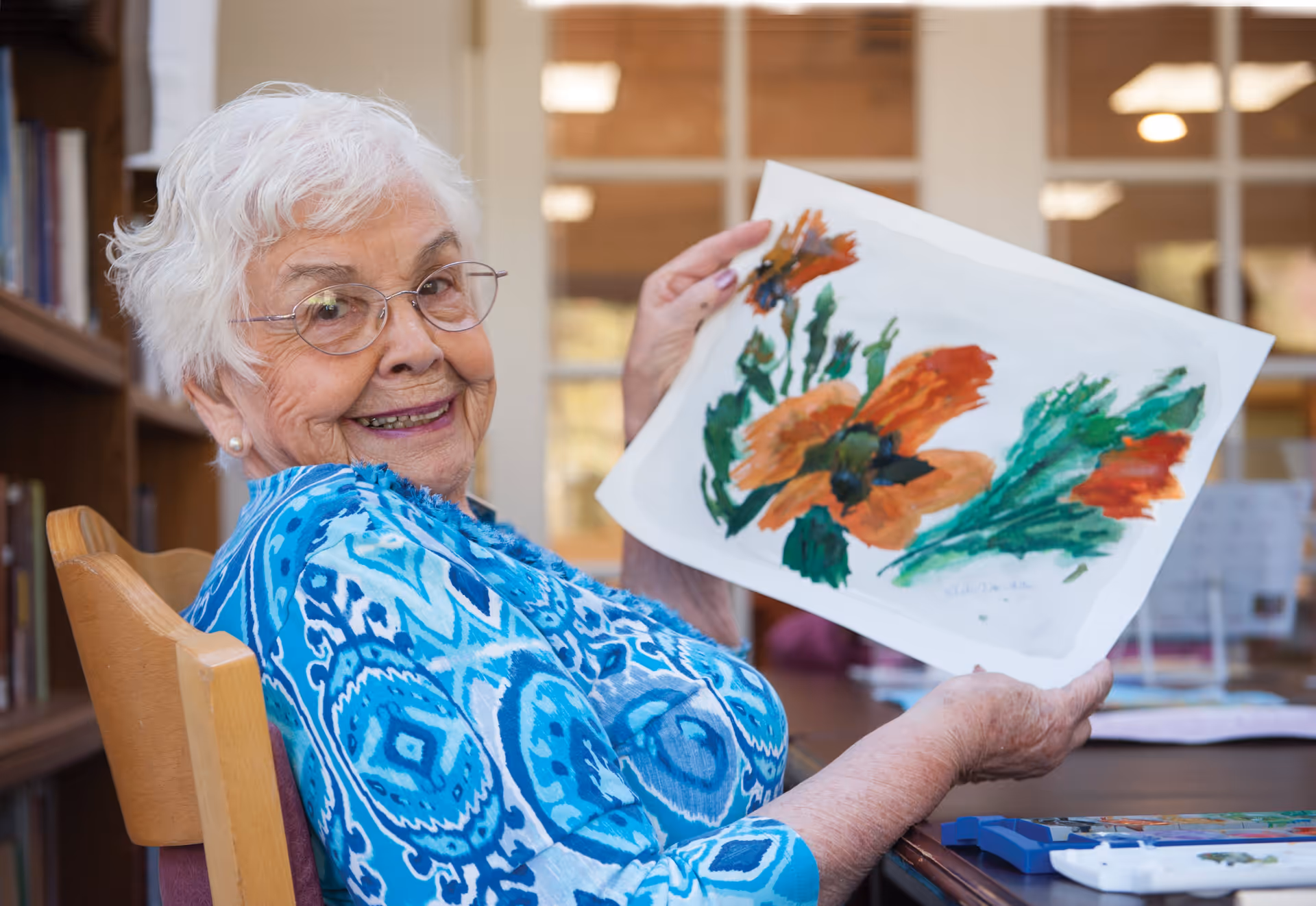 An elderly woman with white hair and glasses, wearing a blue patterned blouse, sitting on a wooden chair and smiling while holding up a painting of orange and green flowers in a well-lit room with bookshelves and large windows in the background.