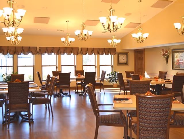 A well-lit dining room with multiple tables and chairs arranged neatly. The room features large windows with valances, several chandeliers hanging from the ceiling, and decorative plants and artwork on the walls.