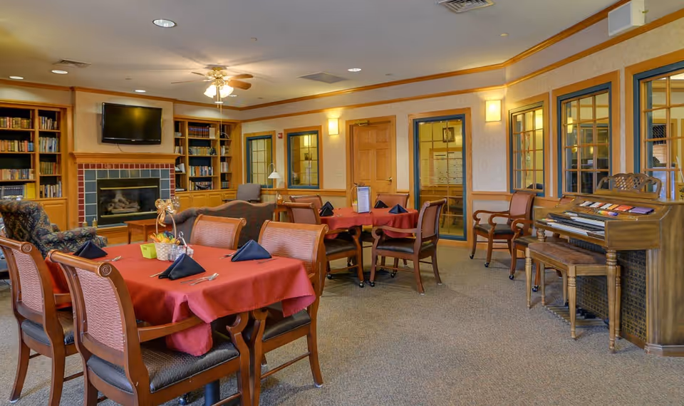 A cozy common area in a senior living facility with tables covered in red tablecloths and set with black napkins and silverware. There are wooden chairs around the tables, a fireplace with a mounted TV above it, bookshelves filled with books, comfortable armchairs, and a piano against the wall. The room has warm lighting and windows with wooden frames.