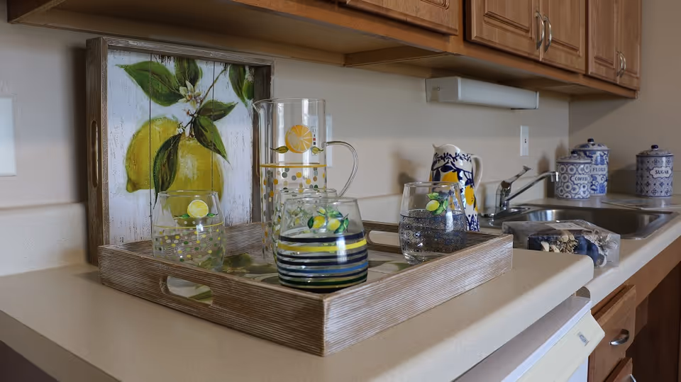 A kitchen countertop with a wooden tray holding a glass pitcher and three glasses decorated with lemon designs. Behind the tray is a decorative wooden panel with a lemon painting. To the right, there is a sink with a faucet, a ceramic pitcher with a blue and yellow pattern, and three blue and white canisters labeled coffee, flour, and sugar.