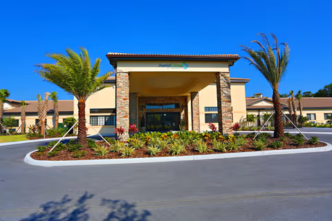 Front exterior view of Inspired Living Sarasota facility with a covered entrance, stone pillars, palm trees, and landscaped plants under a clear blue sky.