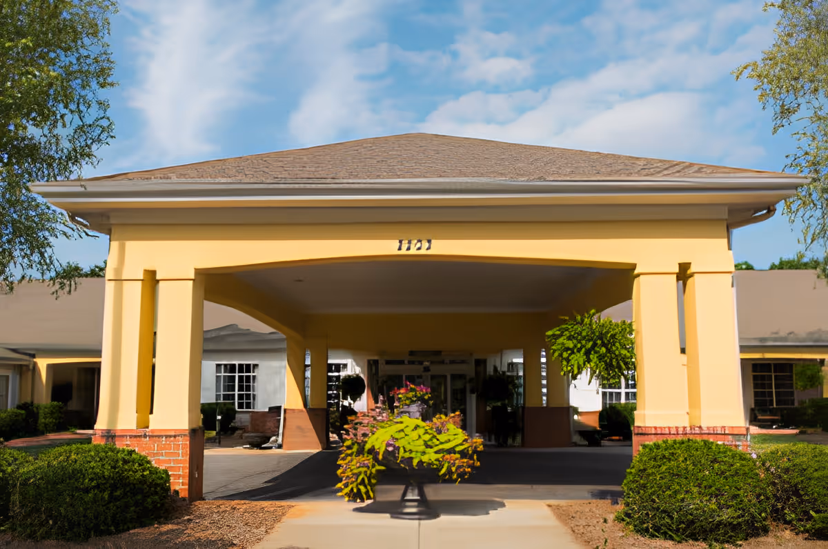 Entrance of a senior living facility with a covered driveway supported by yellow columns and a roof. There are green bushes and plants around the entrance, and a flower arrangement is placed in the center of the driveway. The sky is blue with some clouds.