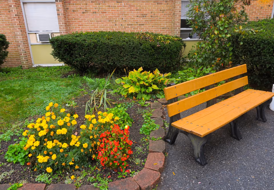 A yellow park bench beside a flowerbed with yellow and red flowers in front of a brick building and shrubs.