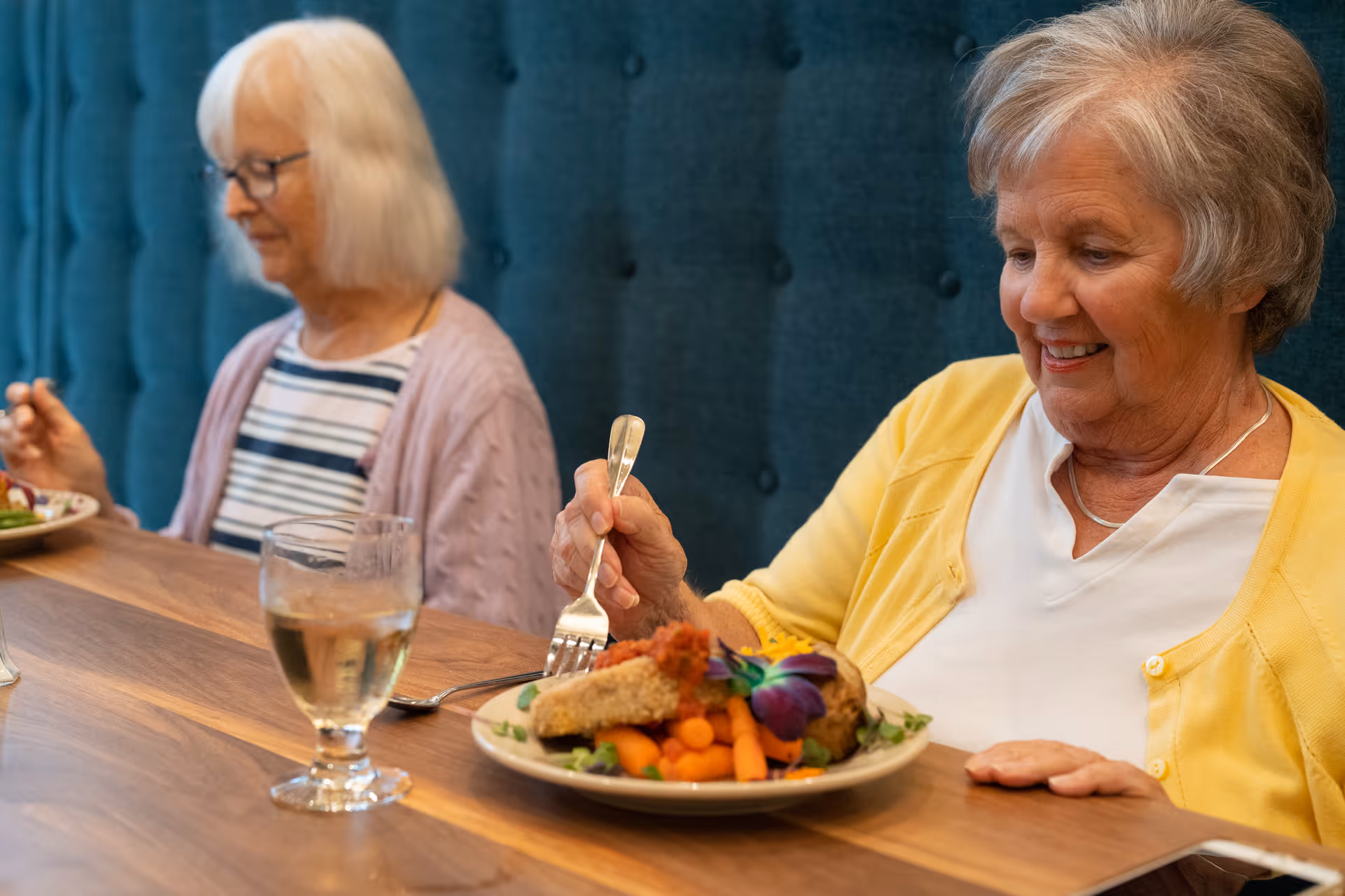 Two elderly women sitting at a wooden table in a dining area. One woman in a yellow cardigan is smiling and holding a fork, about to eat a meal with vegetables and a flower garnish. The other woman with white hair and glasses is sitting beside her, wearing a striped shirt and light purple cardigan. A glass of water is on the table.