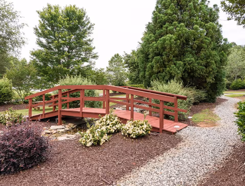 Small red wooden footbridge spanning a landscaped garden with shrubs, trees, and a gravel path.