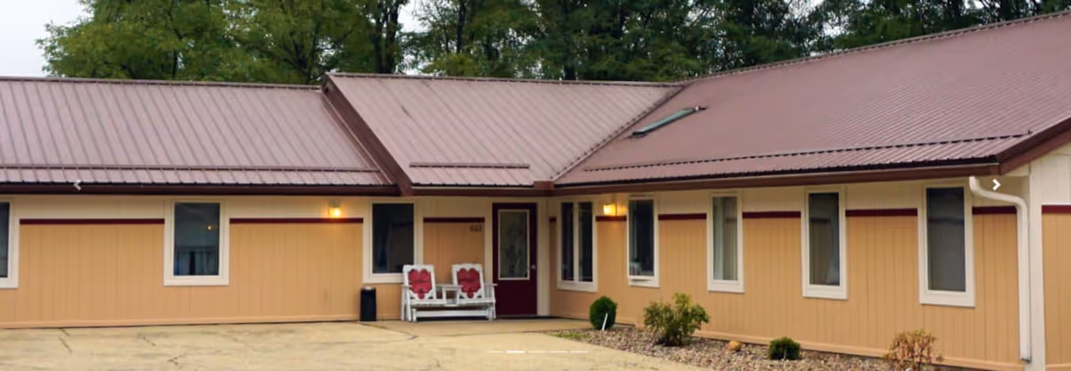 Exterior view of a single-story senior living facility building with a brown metal roof and beige siding. There is a small porch area with two white chairs featuring red cushions near the entrance door. Several windows line the building, and small shrubs are planted along the foundation. Trees are visible in the background.