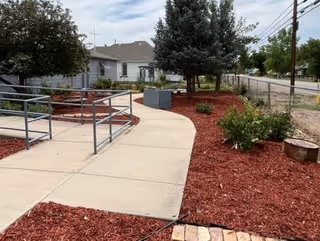 A concrete accessible ramp and walkway with metal railings through red-mulched landscaping leading toward a single-story building and trees.