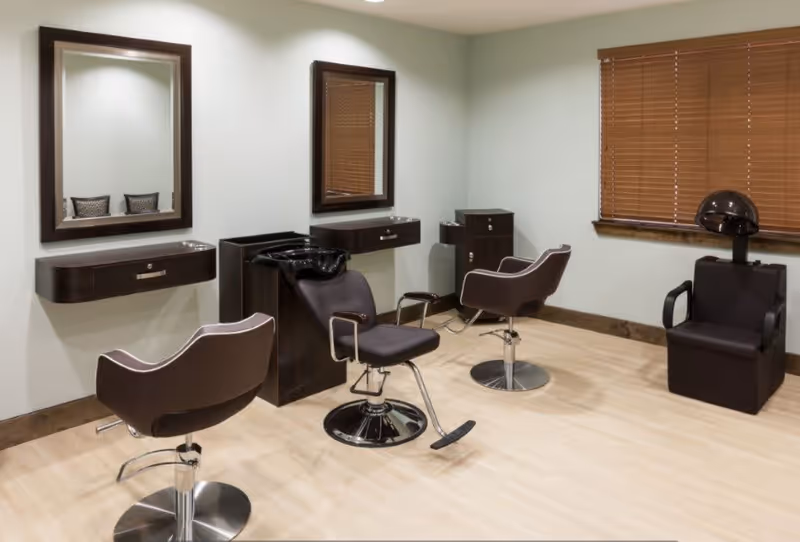 Interior salon room with styling chairs, mirrors, a shampoo sink and a hooded dryer beneath wooden blinds.