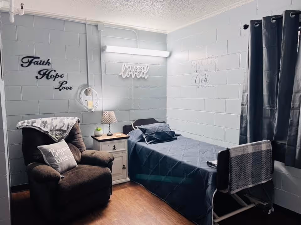 A small, cozy room with a single bed covered in a dark blue quilt and pillows. Next to the bed is a white nightstand with a lamp and a small plant. A comfortable brown recliner chair with a blanket and a pillow that says 'Beyond blessed' is positioned to the left. The walls are painted light gray and decorated with inspirational words such as 'Faith Hope Love' and 'You are so loved'. A window with dark curtains is on the right side of the room.