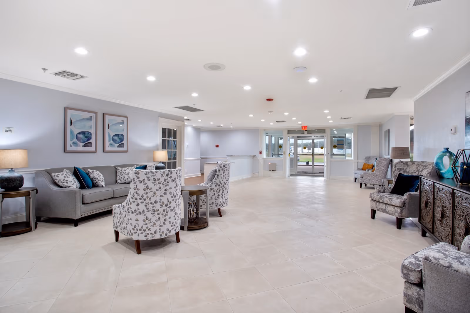 A spacious and well-lit senior living facility lobby with light-colored tiled flooring and soft gray walls. The seating area includes a gray sofa with patterned and solid blue cushions, two patterned armchairs, and additional armchairs along the walls. There are side tables with lamps and decorative items, and framed abstract artwork hangs on the wall. Large glass doors lead to the outside, allowing natural light to enter the space.