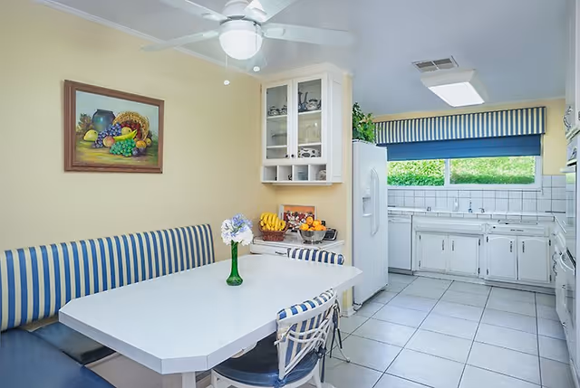 Bright kitchen dining nook with a white table, blue-striped banquette and chairs, a small vase of flowers, and a view into a white tiled kitchen.