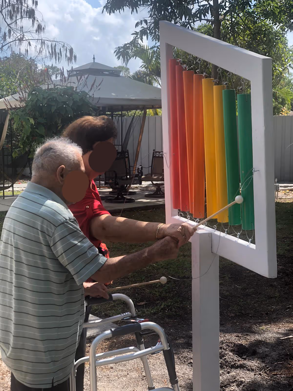 An elderly man using a walker and a woman standing beside him are playing a large outdoor musical instrument with colorful vertical tubes in a garden area. There is a gazebo and some chairs in the background, along with trees and a fence.