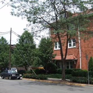 Street view of a red brick building partially obscured by trees and bushes, with a parked black vehicle on the side of the road and power lines overhead.