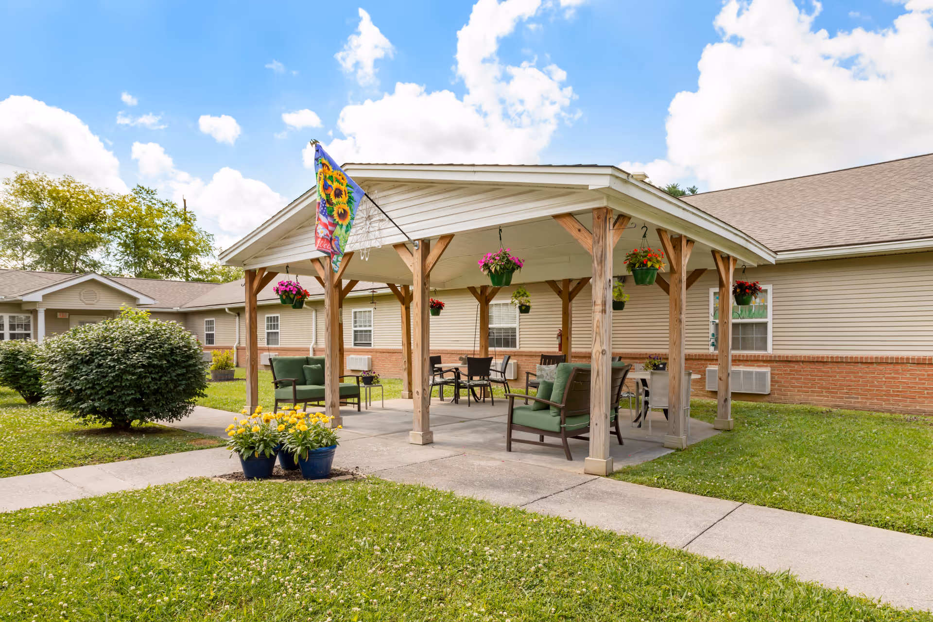 Outdoor covered seating area with wooden posts and a white roof, featuring green cushioned chairs, tables, hanging flower pots, and a colorful flag with sunflowers. The area is surrounded by grass, bushes, and a beige building with windows in the background under a partly cloudy blue sky.