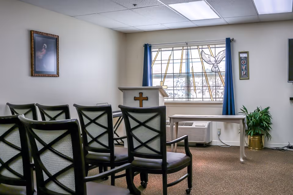 A small chapel or prayer room with several chairs arranged in rows facing a podium with a cross on it. There is a stained glass window with a dove design behind the podium, blue curtains on either side, a framed picture on the wall, a small table, a potted plant, and an air conditioning unit beneath the window.