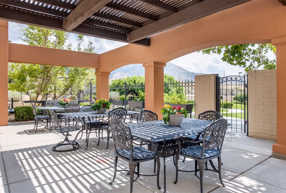 Outdoor covered patio area with metal tables and chairs featuring decorative backs and cushions. Each table has a flower pot centerpiece. The patio has a pergola-style roof casting striped shadows on the ground. Beyond the patio, there is a black metal gate and lush greenery with trees and bushes, and mountains are visible in the background under a partly cloudy sky.