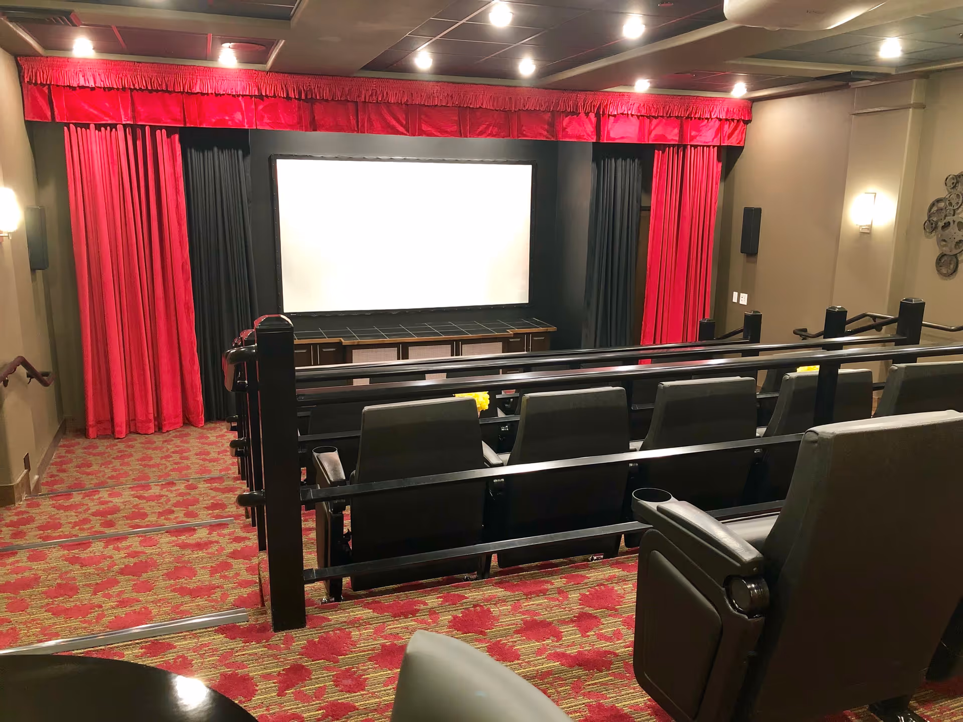 Interior view of a small theater room with a large white screen framed by red and black curtains. The room has tiered seating with comfortable, dark-colored chairs and a red patterned carpet. The walls are beige with modern light fixtures and decorative film reels on one side.