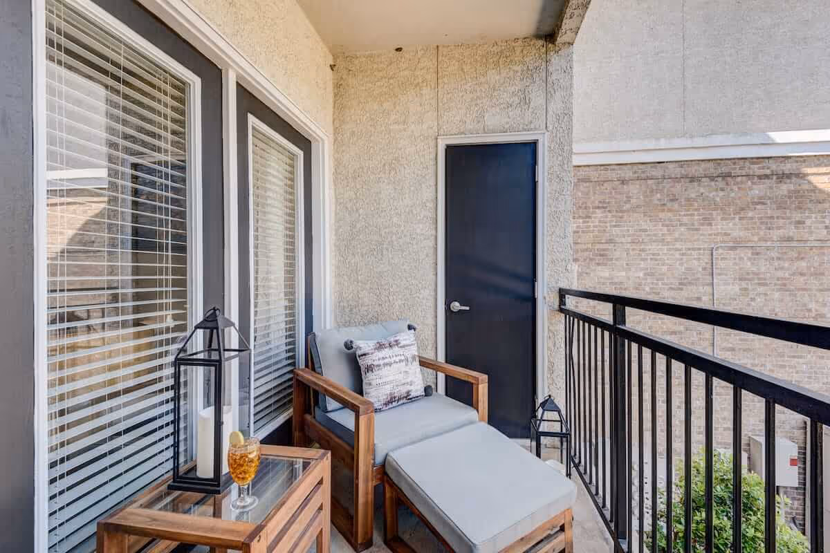 A small balcony with a cushioned wooden lounge chair and matching ottoman, a side table with a glass of iced tea and a decorative lantern, black metal railing, and a closed black door. The balcony overlooks a brick wall and some greenery.