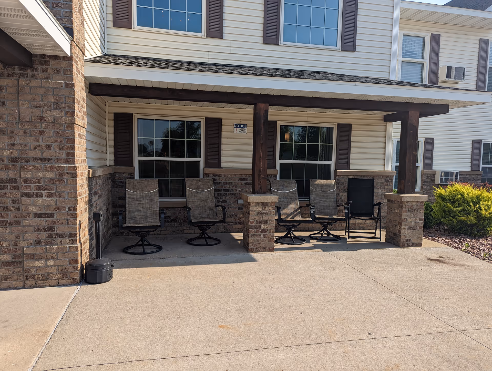Outdoor covered patio area with four chairs arranged in front of two windows on a brick and siding building exterior. There is a concrete floor and some landscaping with bushes visible to the right.