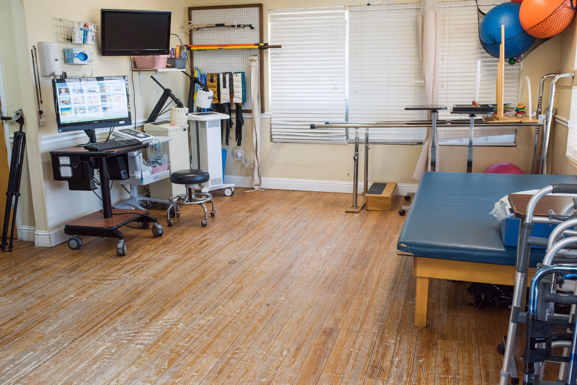 A rehabilitation therapy room with wooden flooring, a blue therapy table, parallel bars for walking exercises, exercise balls, a walker, a computer workstation on wheels, and various therapy equipment hanging on the wall and stored around the room. The room has windows with blinds partially closed.