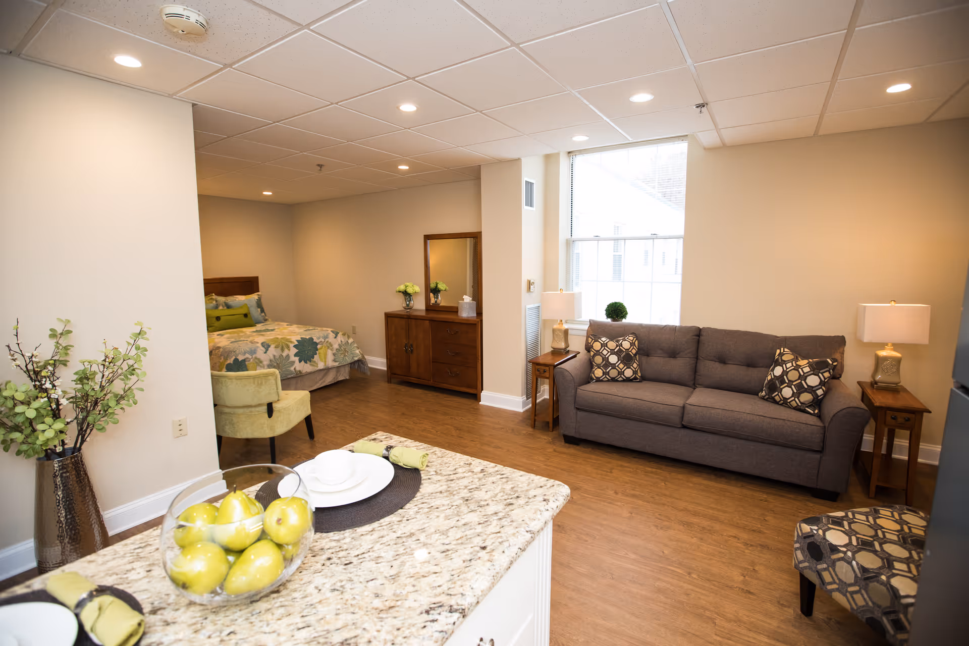 A bright and cozy studio apartment in a senior living facility featuring a bed with floral bedding, a wooden dresser with a mirror, a gray sofa with patterned pillows, two side tables with lamps, a patterned armchair, and a granite countertop with a bowl of green apples and place settings. The room has wooden flooring and a large window letting in natural light.
