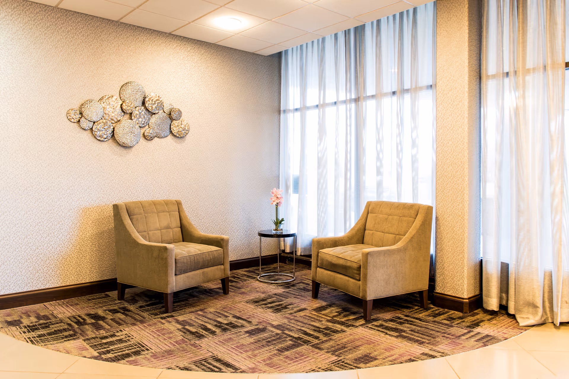 A cozy sitting area with two beige armchairs facing each other, separated by a small round table with a pink flower in a vase. The area has patterned carpet flooring and large windows with sheer white curtains allowing natural light to fill the space. A decorative metal wall art piece is mounted on the wall behind one of the chairs.