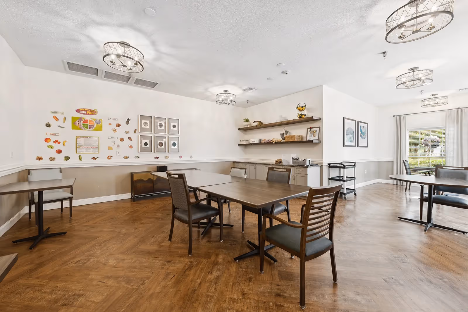 A bright dining room with wooden floors and several tables and chairs arranged for seating. The walls are decorated with educational posters about nutrition and framed pictures. There are modern light fixtures on the ceiling and a window with curtains letting in natural light. Shelves on one wall hold decorative items and kitchen appliances.