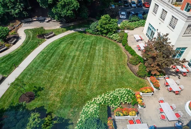 Aerial view of a well-maintained outdoor garden area with a large green lawn, curved walking paths, benches, colorful flower beds, and patio tables with red chairs next to a white building. Several parked cars are visible in the background.